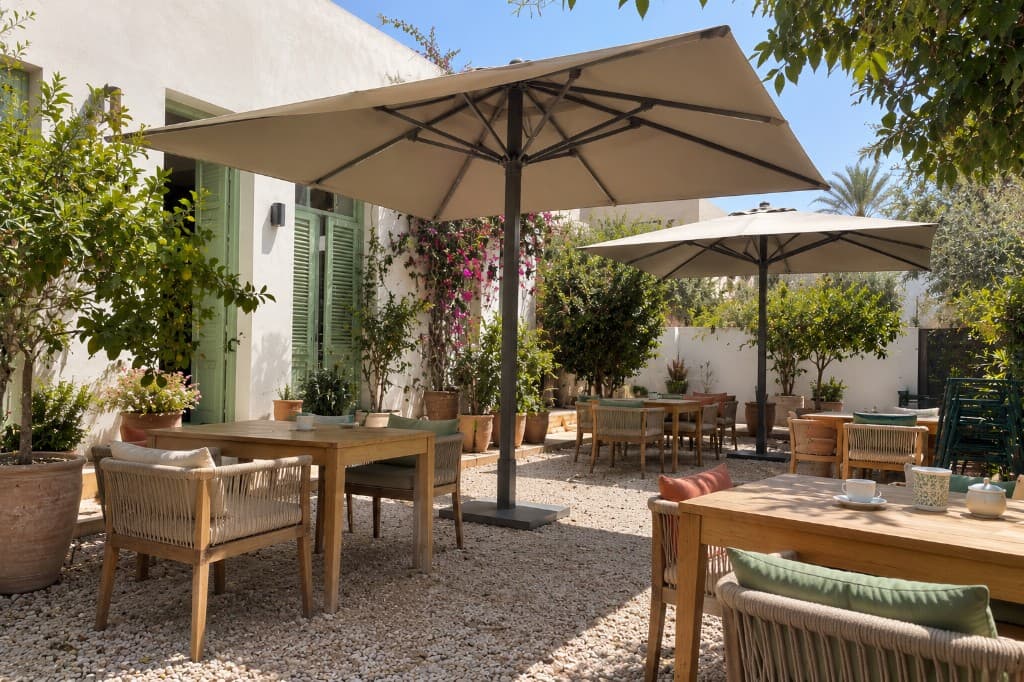 Deux parasols à mât central, toiles carrées claires, au-dessus de tables dans une cour.