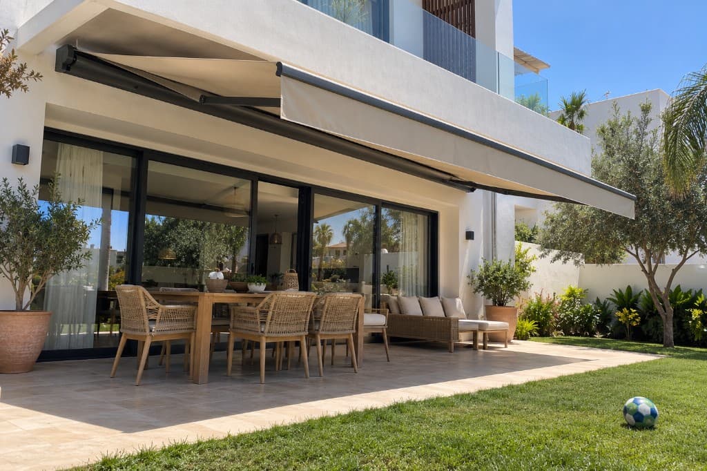 Store banne beige déployé devant une baie de villa, bras articulés visibles, table en terrasse.