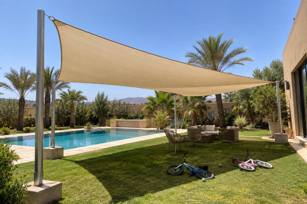 Voile d'ombrage beige tendue sur des mâts métalliques ancrés dans des plots, au-dessus d'un jardin.